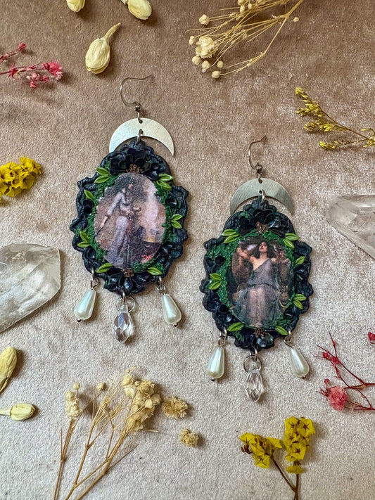 Pair of earrings with a central figure surrounded by greenery and crystals on a textured surface with dried flowers.